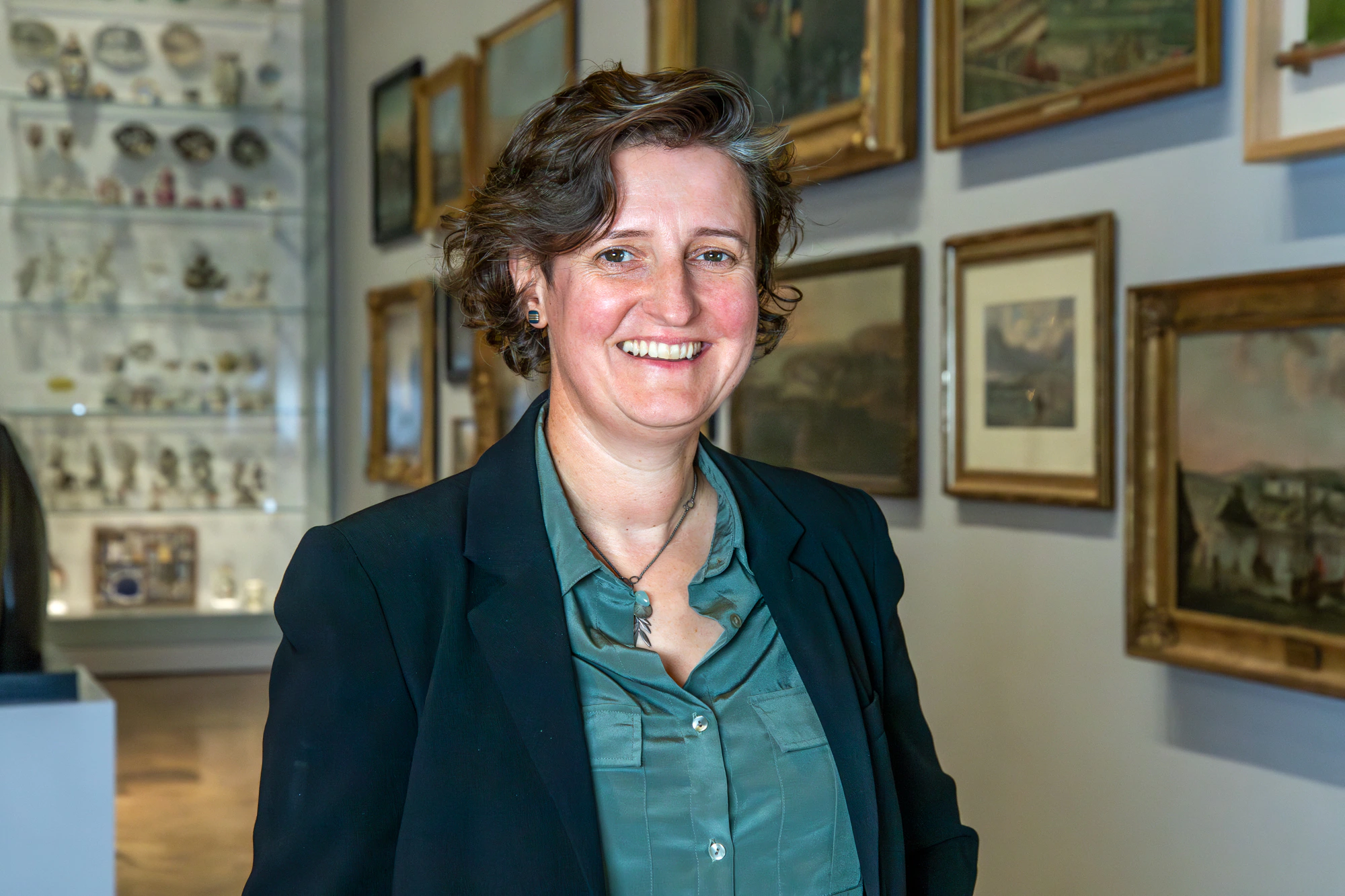 A person with short, wavy brown hair, wearing a dark blazer and green shirt, smiles while standing in an art gallery with framed paintings and glass display cases in the background.