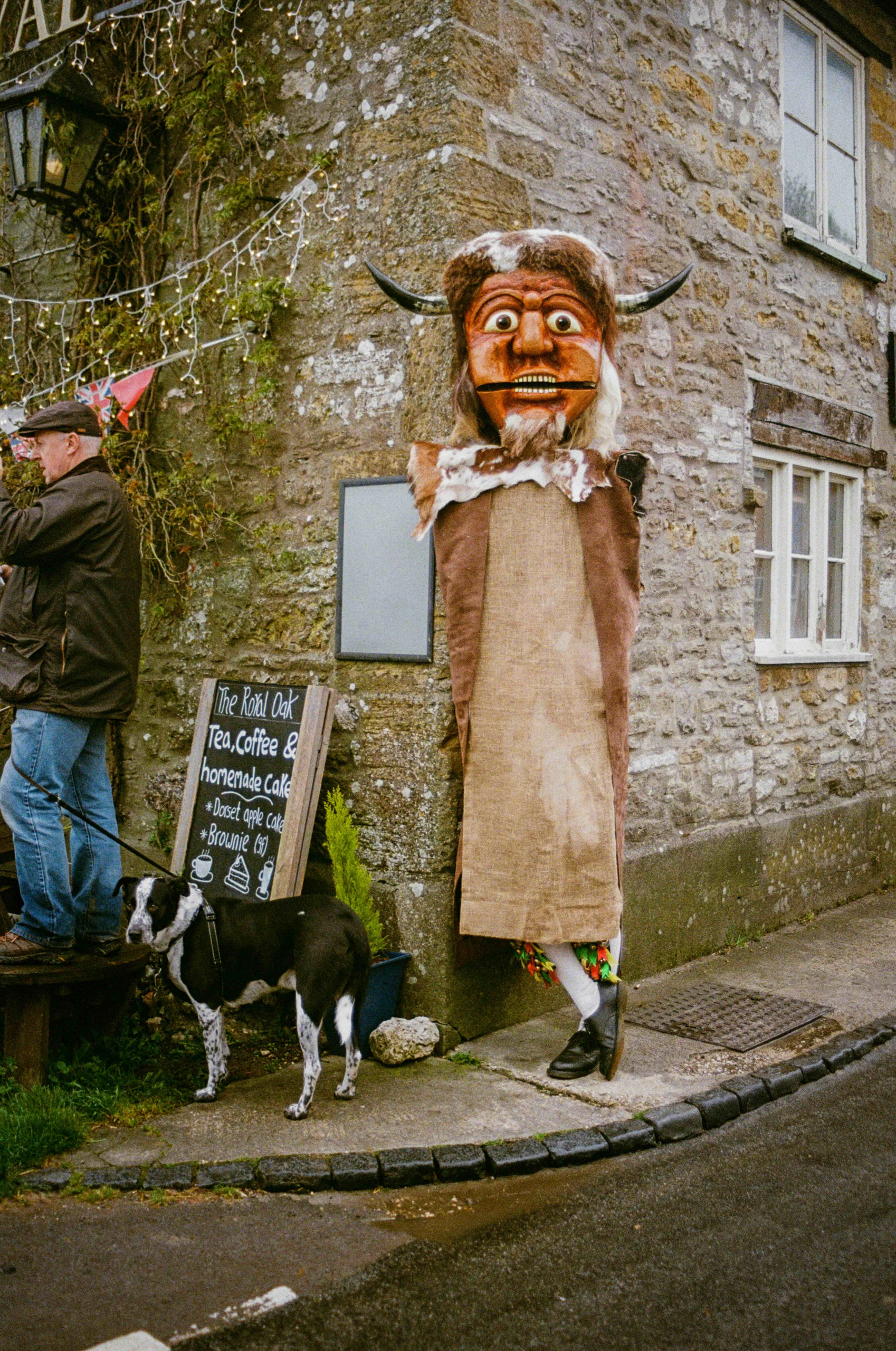 A large costumed figure with a horned mask and brown cloak stands by a stone building, next to a chalkboard advertising tea and coffee. A black and white dog is tied nearby, and a person stands on a step to the left.