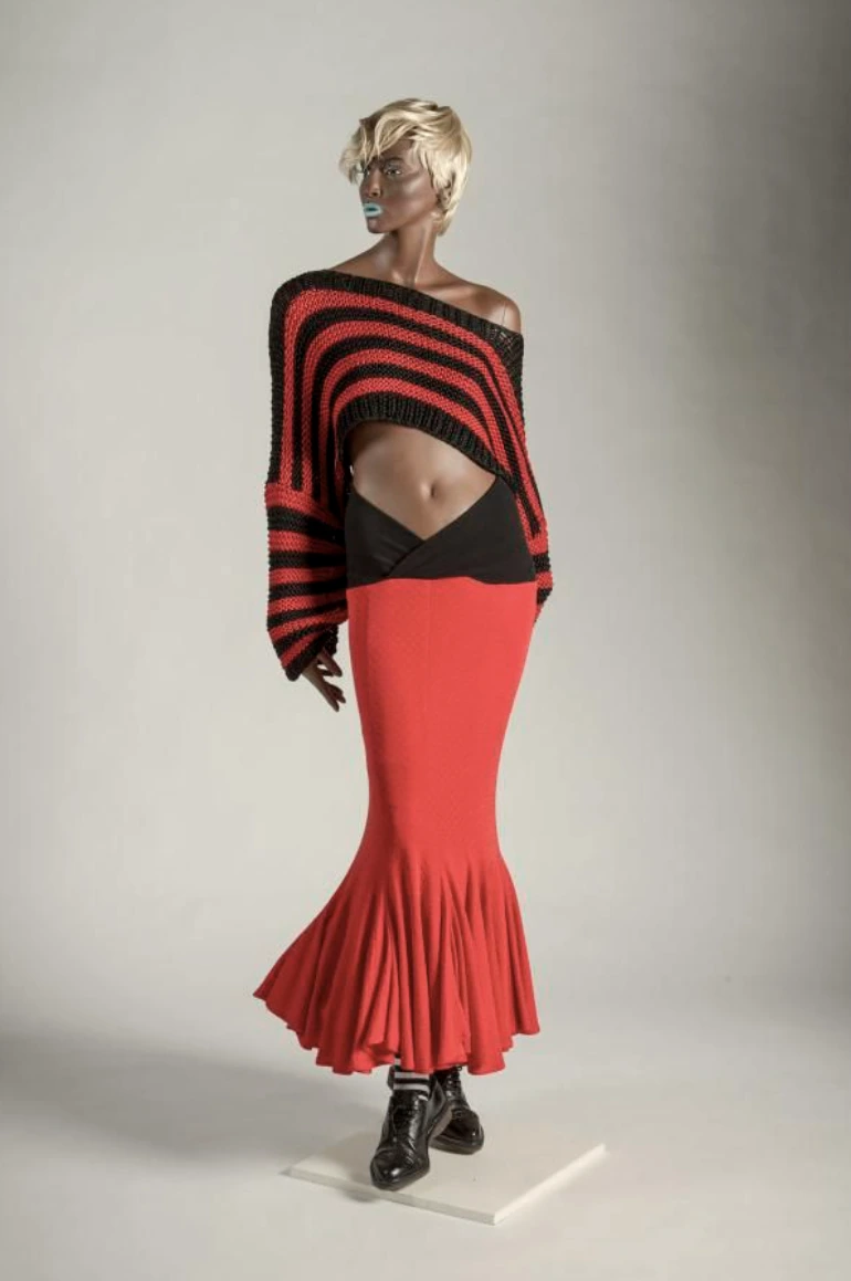 A mannequin wears a red flared skirt, black boots, and an off-the-shoulder black and red striped sweater that reveals the midriff. The mannequin has short blonde hair and sunglasses against a plain background.