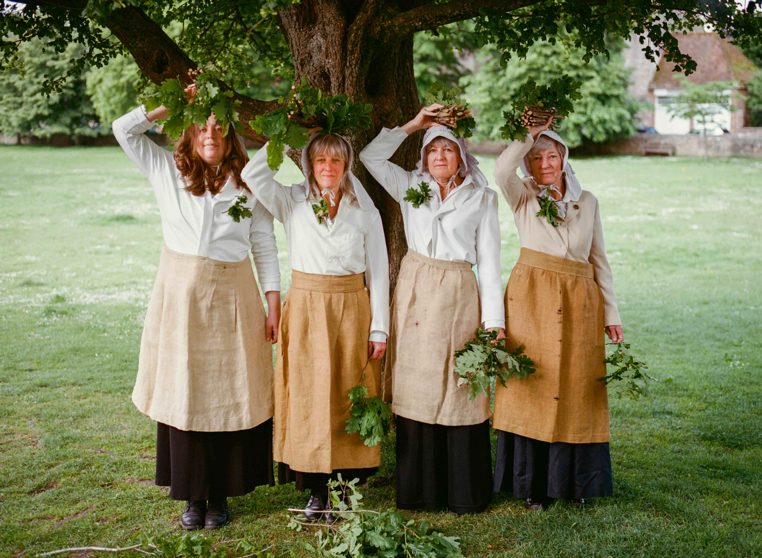 Four women standing under a tree in a grassy field, wearing white shirts, tan aprons, and long dark skirts, holding green branches and leaves, with some branches held above their heads.