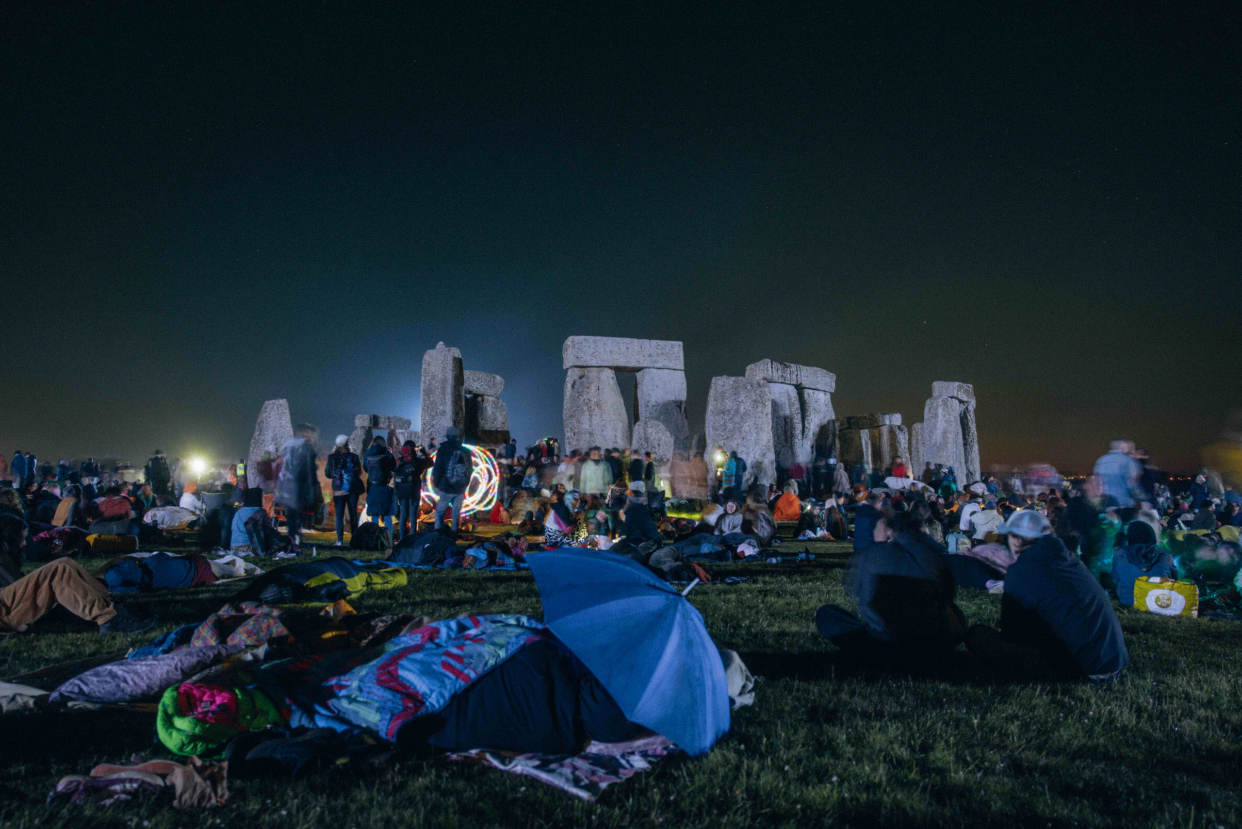 A large crowd gathers and camps on the grass at night in front of Stonehenge, under a dark sky, with some people sitting, lying down, and socializing near the ancient stone monument.