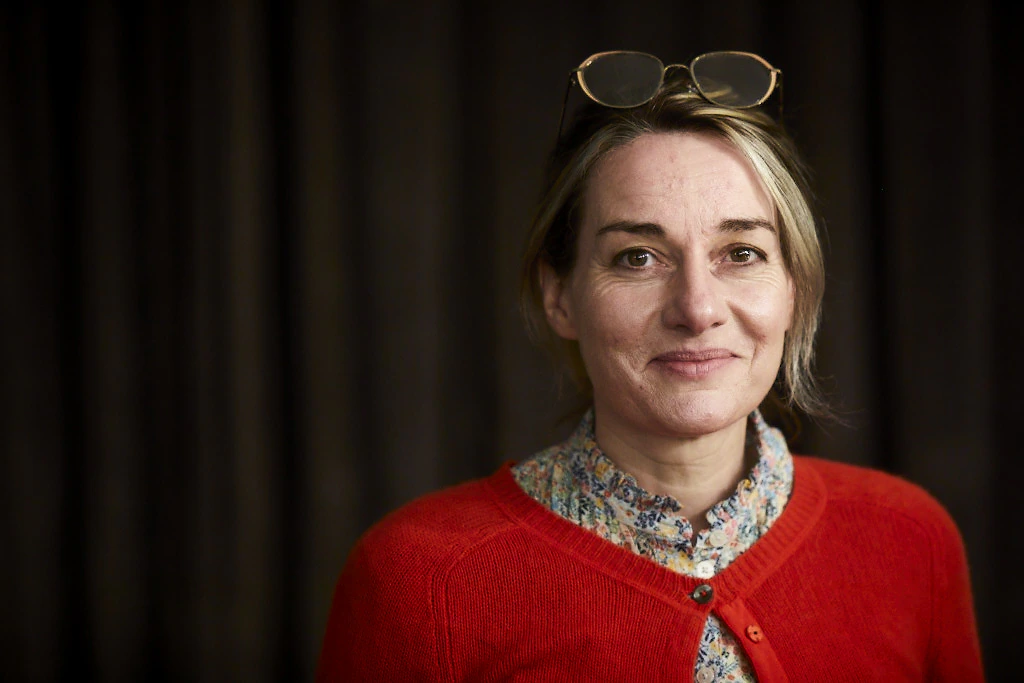 A woman with light hair wears glasses on her head, a floral shirt, and a bright red sweater, smiling softly while standing in front of a dark, blurred background.