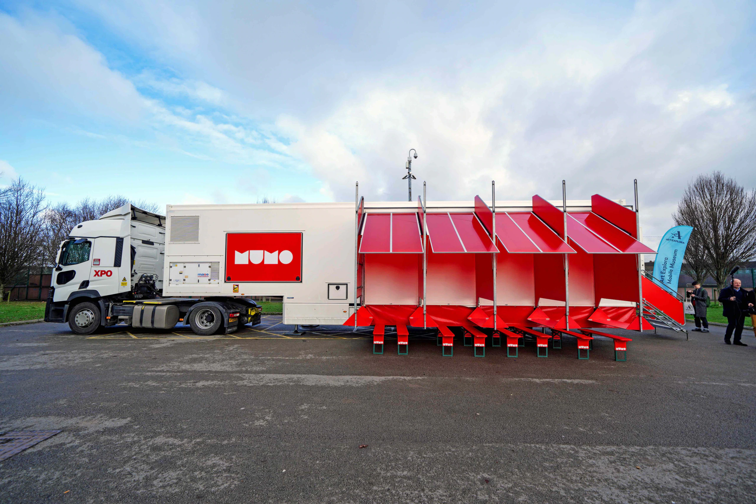 A large white truck with a red MUWO logo is parked, attached to a mobile structure featuring five bright red, angled modular seating units, set outdoors on a paved area with trees and cloudy sky in the background.