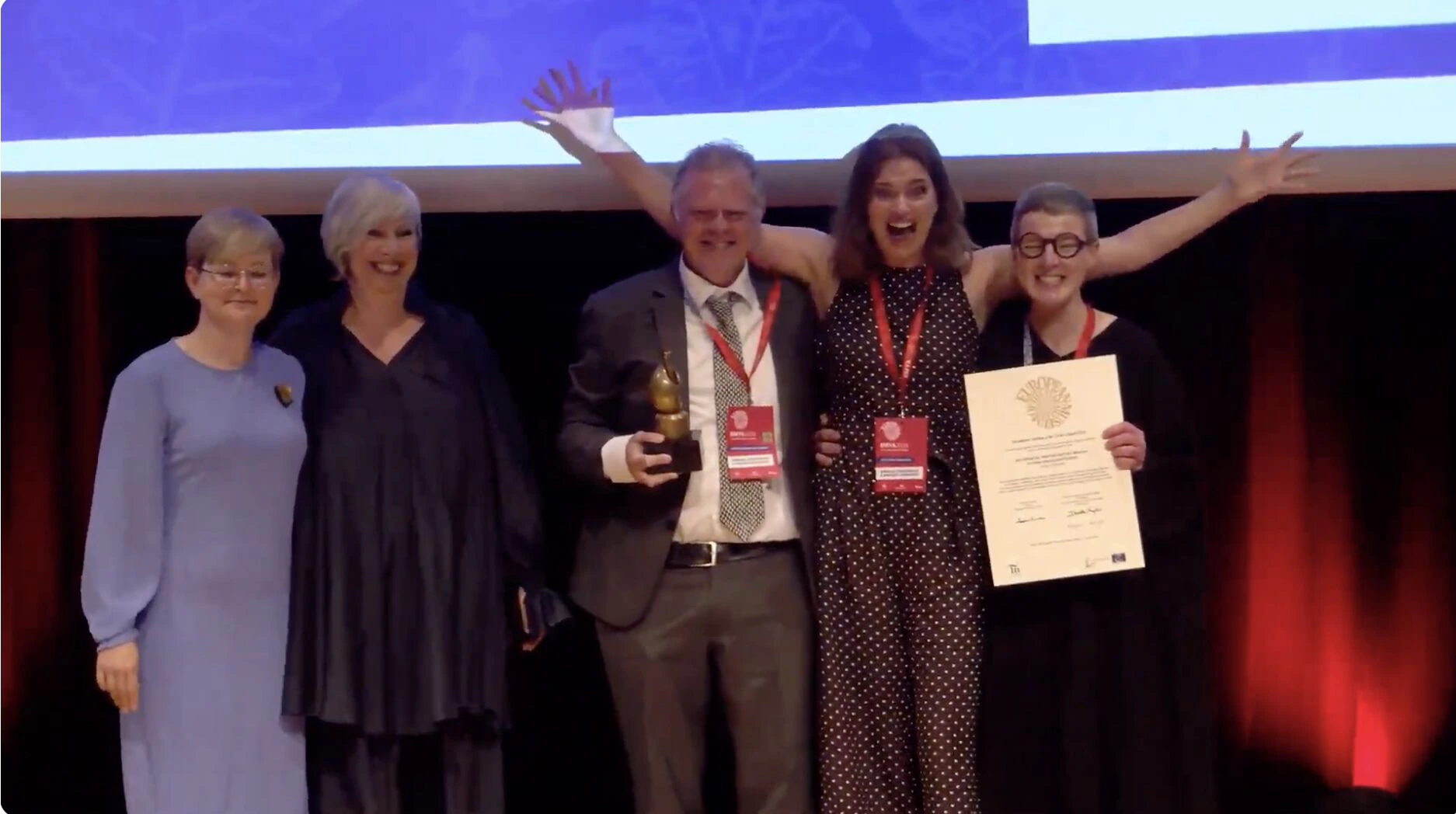 Five people stand on stage at an award ceremony. One man in a suit holds a trophy, while a woman beside him celebrates with arms raised. Another woman holds a large certificate. Everyone is smiling under stage lights.