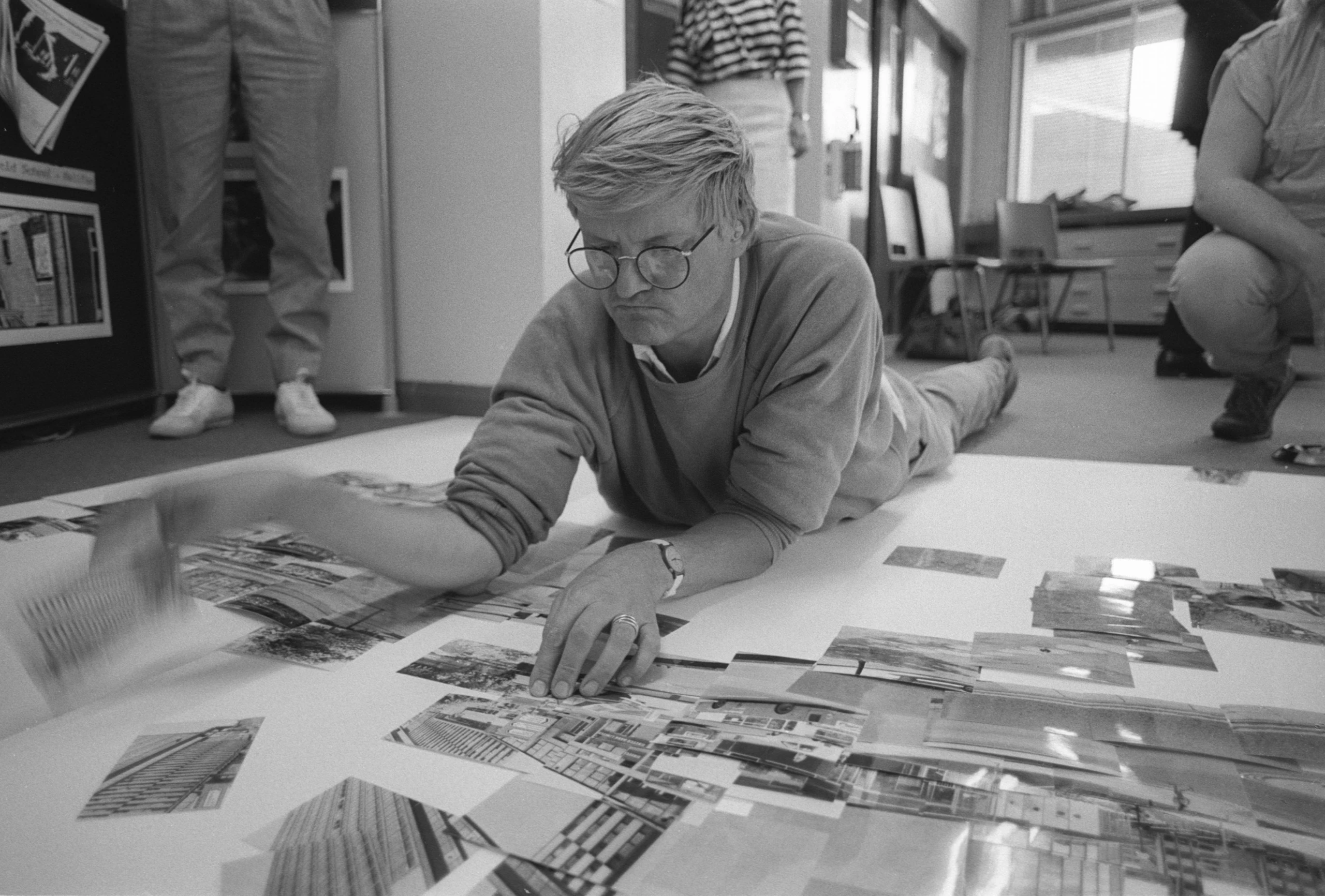 A person with glasses lies on the floor, arranging black-and-white photo prints on a large white sheet. Three other people stand or sit nearby in a bright, modern room with windows and chairs.