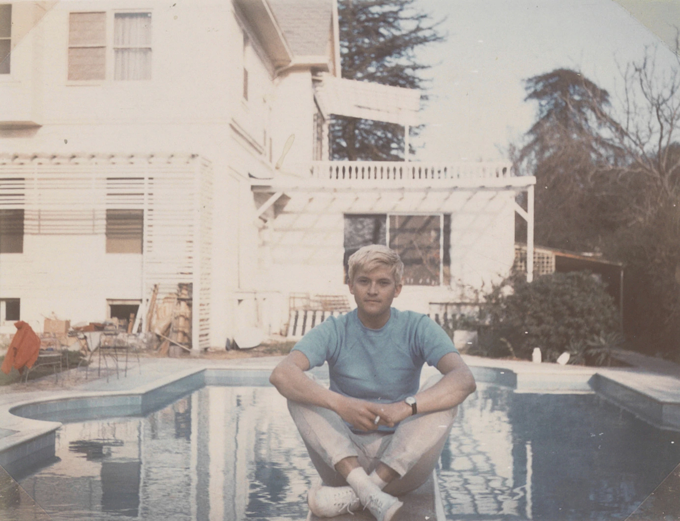 A young man with blond hair, wearing a light blue shirt and light pants, sits cross-legged at the edge of a backyard pool in front of a large white house, with patio furniture and trees in the background.