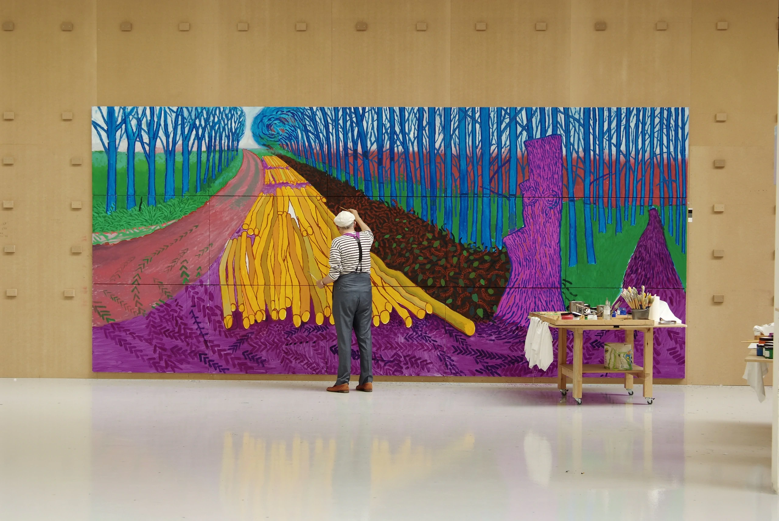 An artist wearing a hat and overalls paints a large, colorful landscape mural featuring a vibrant road, fields, and trees inside a spacious, modern studio with art supplies on a nearby table.