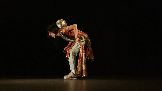 A person in a colorful outfit and sneakers bends forward on stage, balancing a small globe on their upper back against a dark background.