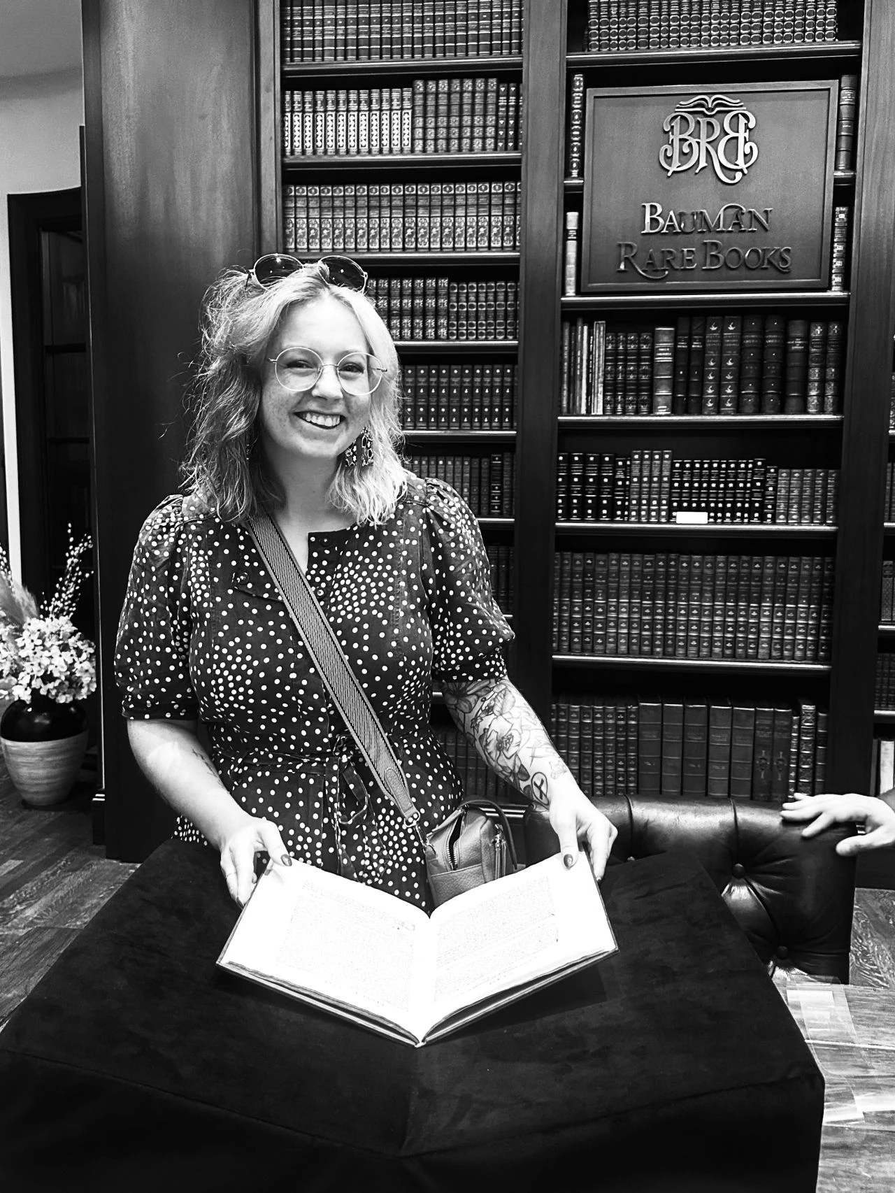 A smiling woman with glasses and shoulder-length hair stands in front of a bookshelf labeled Bauman Rare Books, holding an open book on a display stand in a library or bookstore.