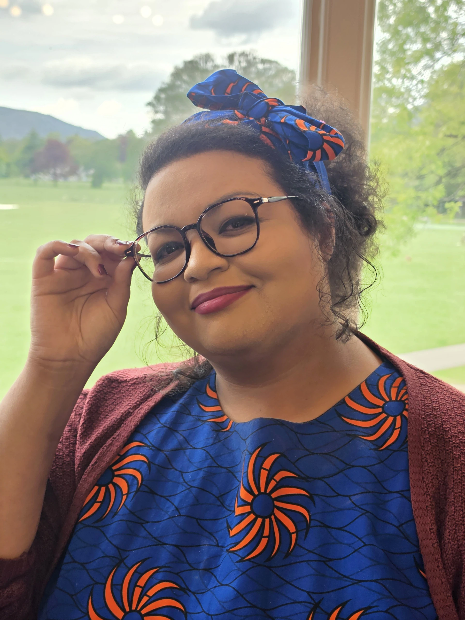 A smiling person wearing glasses, a patterned blue and orange dress, a matching headband, and a maroon sweater poses indoors near a window with a green outdoor landscape in the background.