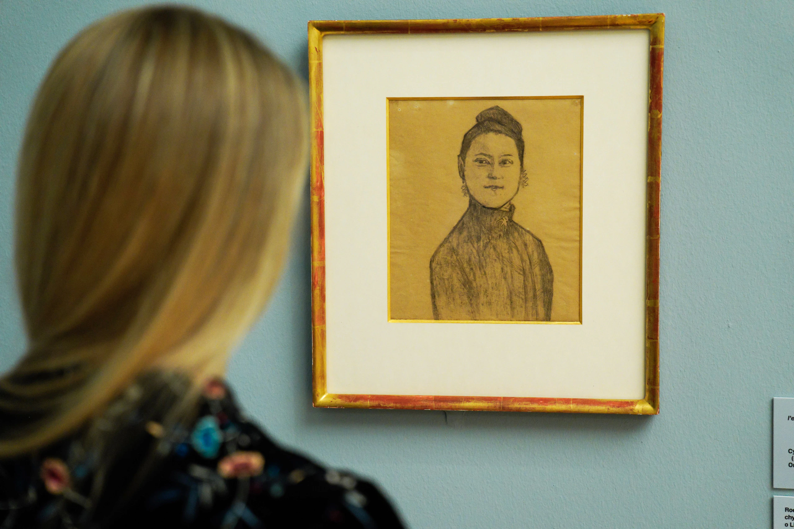 A woman with long blonde hair looks at a framed pencil sketch of a person with dark hair and a high collar, displayed on a light blue wall in an art gallery.