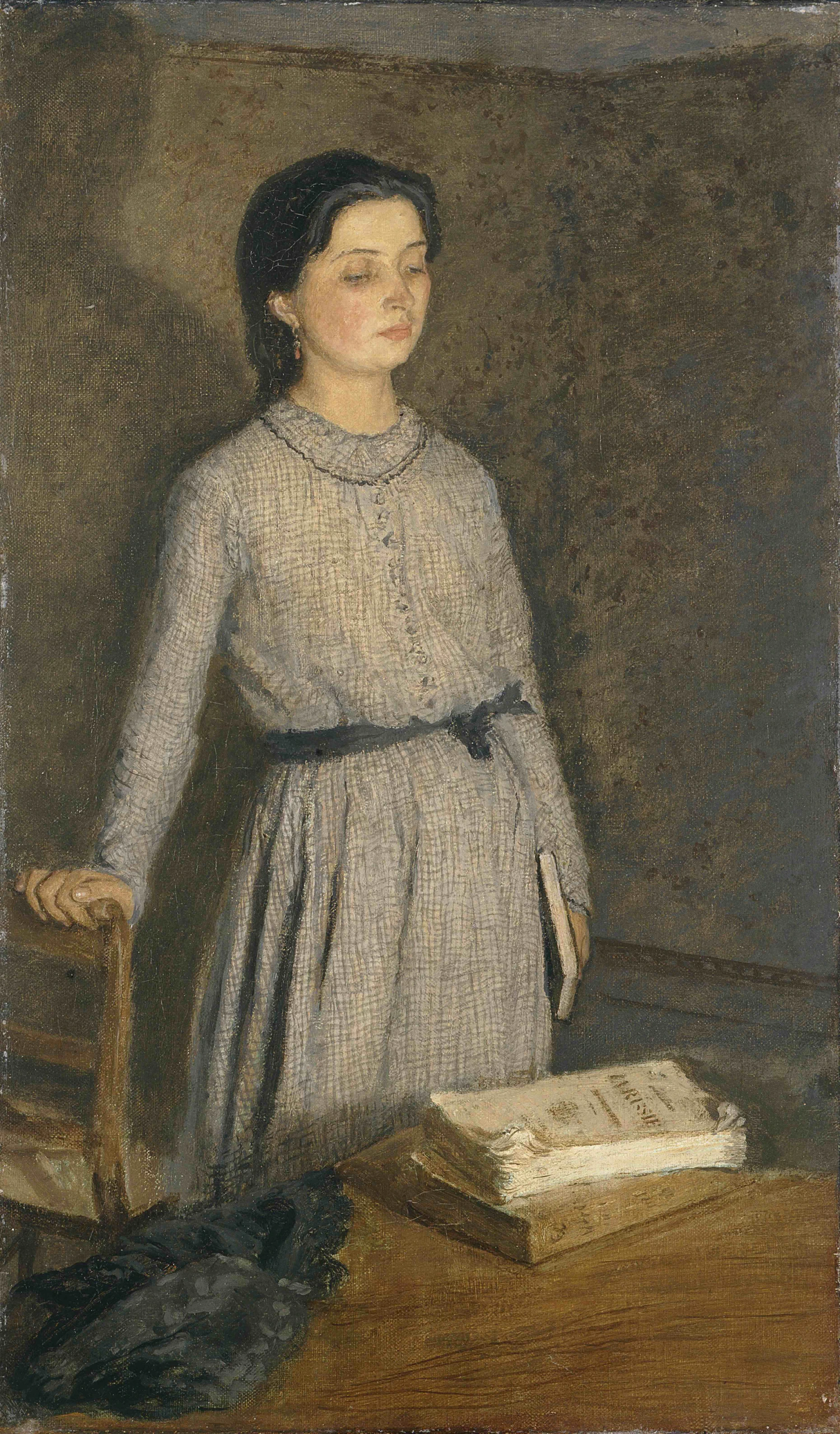 A young girl in a simple gray dress stands beside a wooden table, holding a closed book in one hand. On the table are more books and a dark hat. The background is plain and muted.