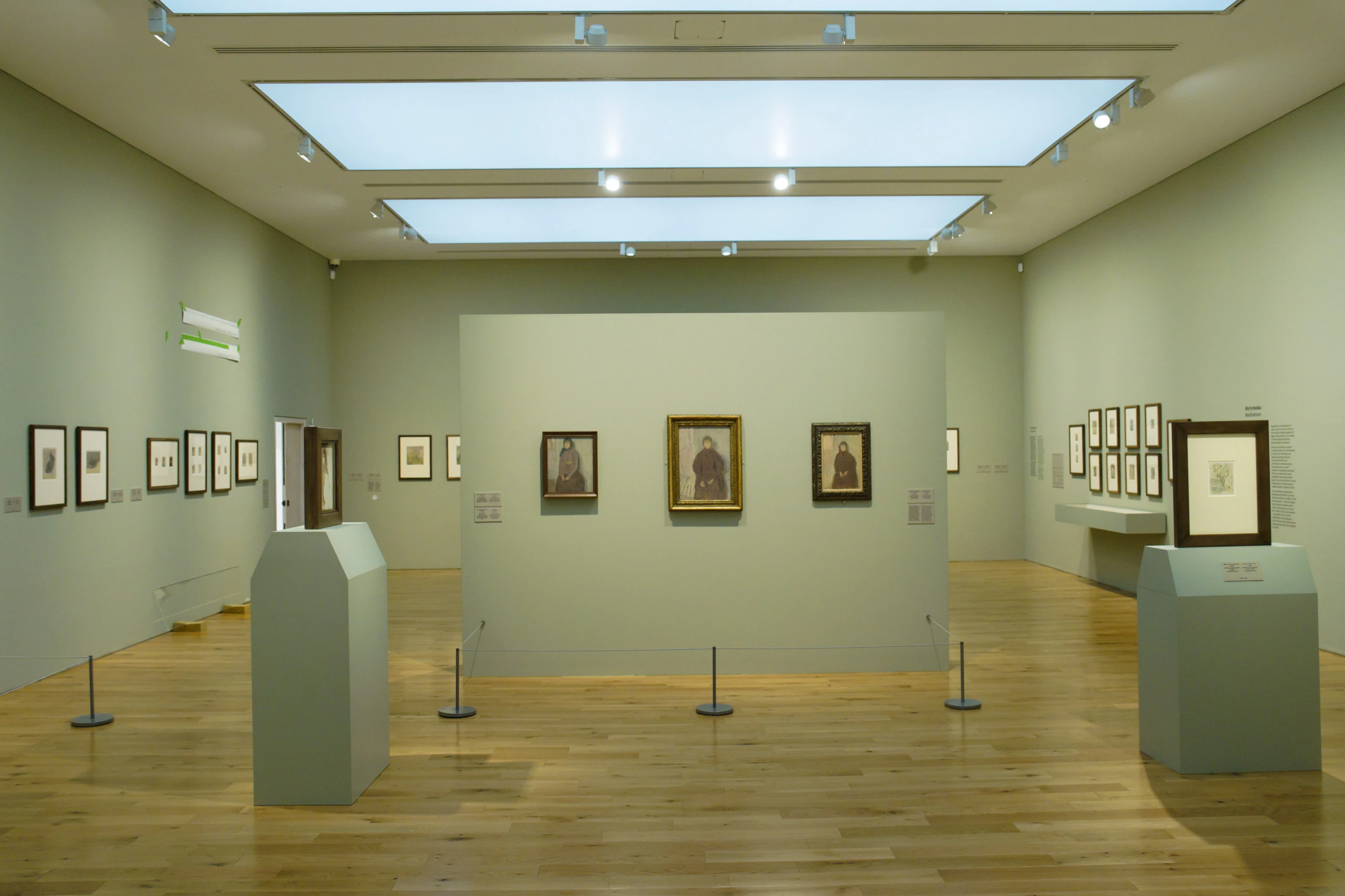 A spacious, modern art gallery with light wood floors, pale green walls, and framed artwork displayed on walls and freestanding panels under bright ceiling lights.