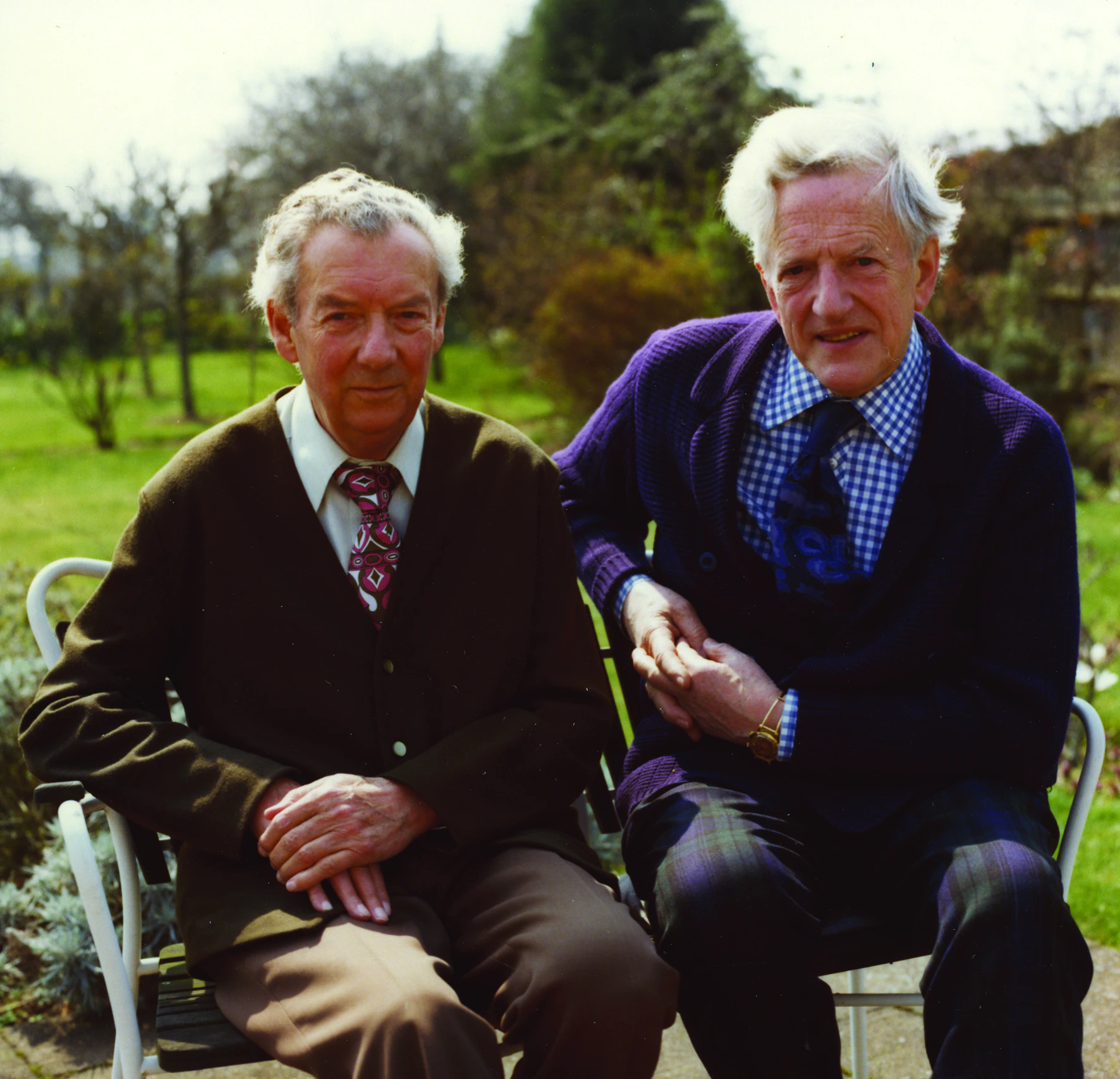 Two elderly men sit closely together on garden chairs outdoors, both wearing cardigans and ties. They are smiling slightly, with trees and greenery in the blurred background on a sunny day.