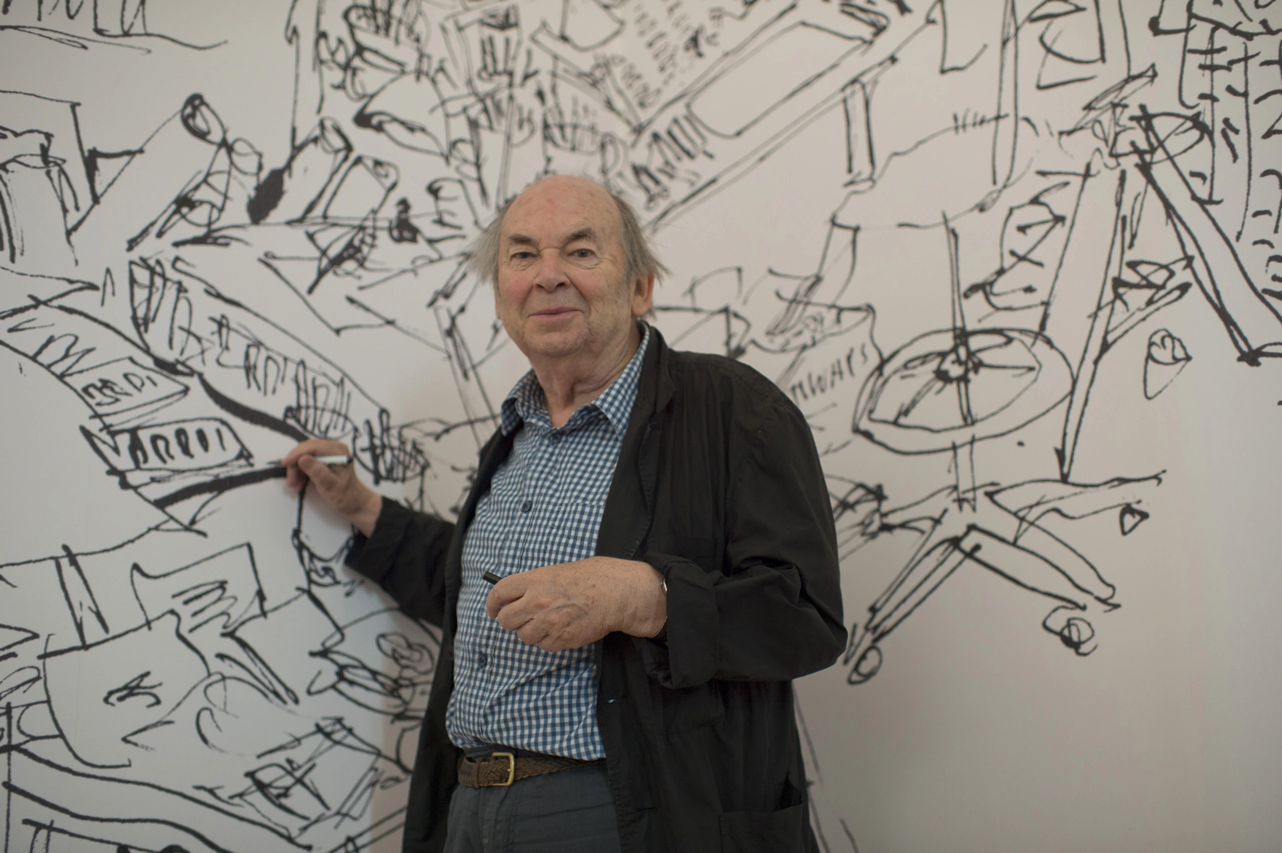 An older man with gray hair and a checkered shirt stands in front of a large wall covered in black-and-white doodles, holding a marker as if drawing.