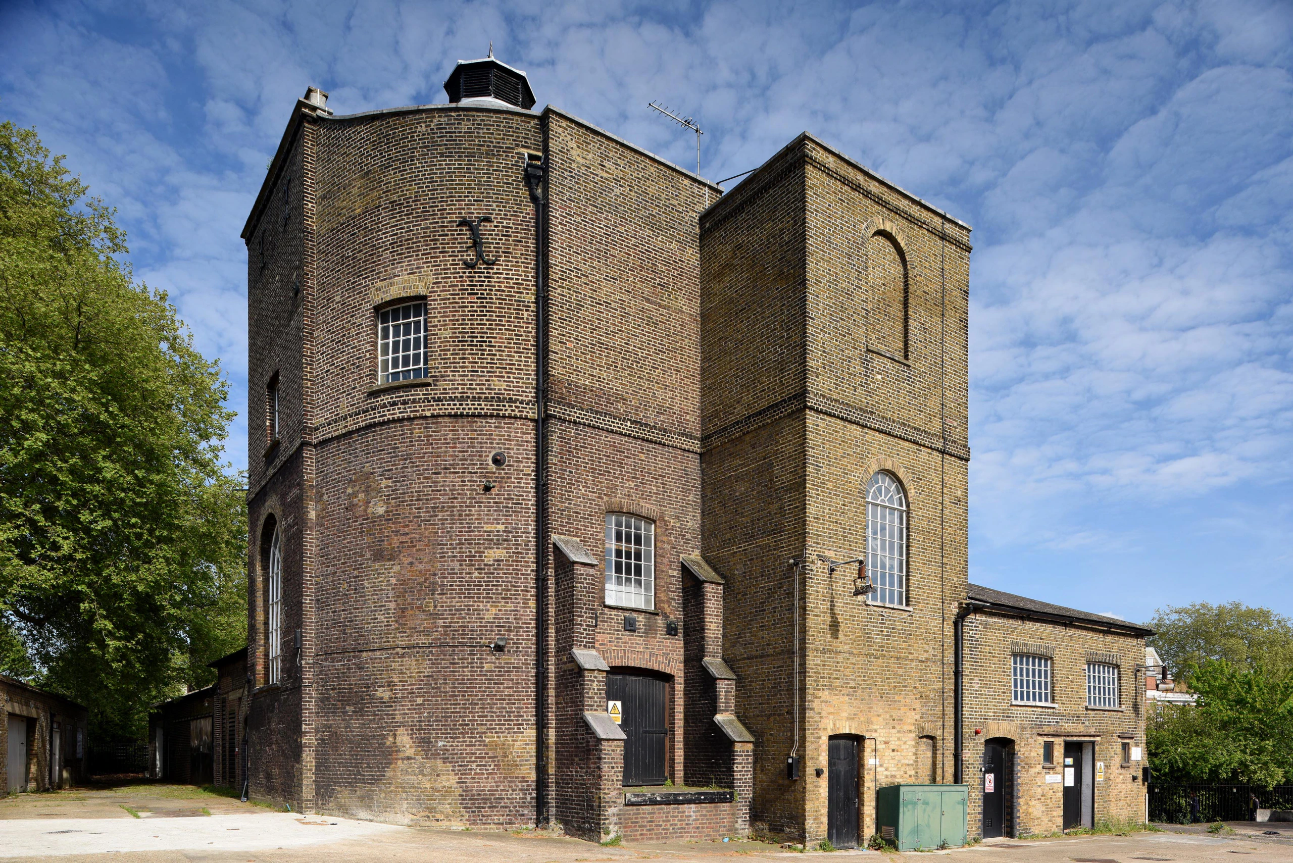 A historic, brick building with tall, arched windows and two main towers stands under a blue sky with scattered clouds. Green trees border the site and a utility box is visible in front of the structure.