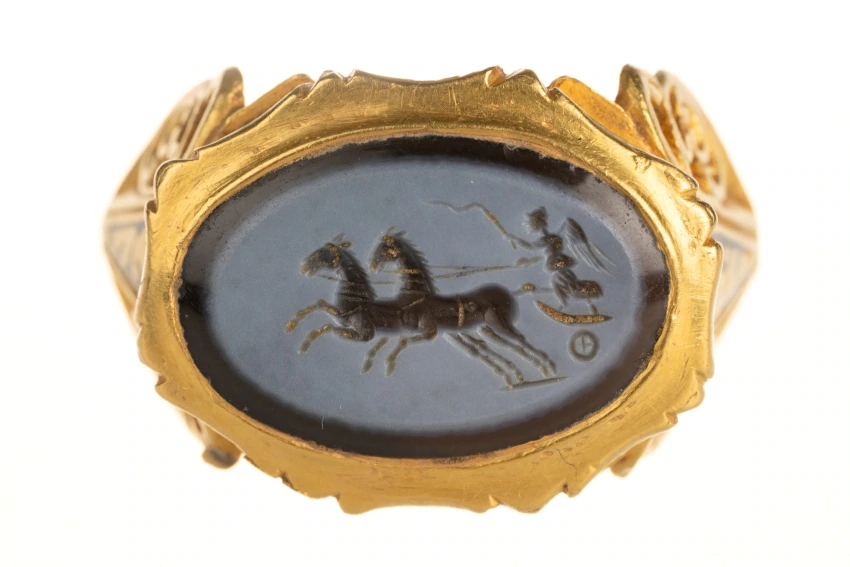 Gold ring with an engraved black oval stone depicting a chariot scene with two horses and a driver, set in an ornate, decorative gold band.