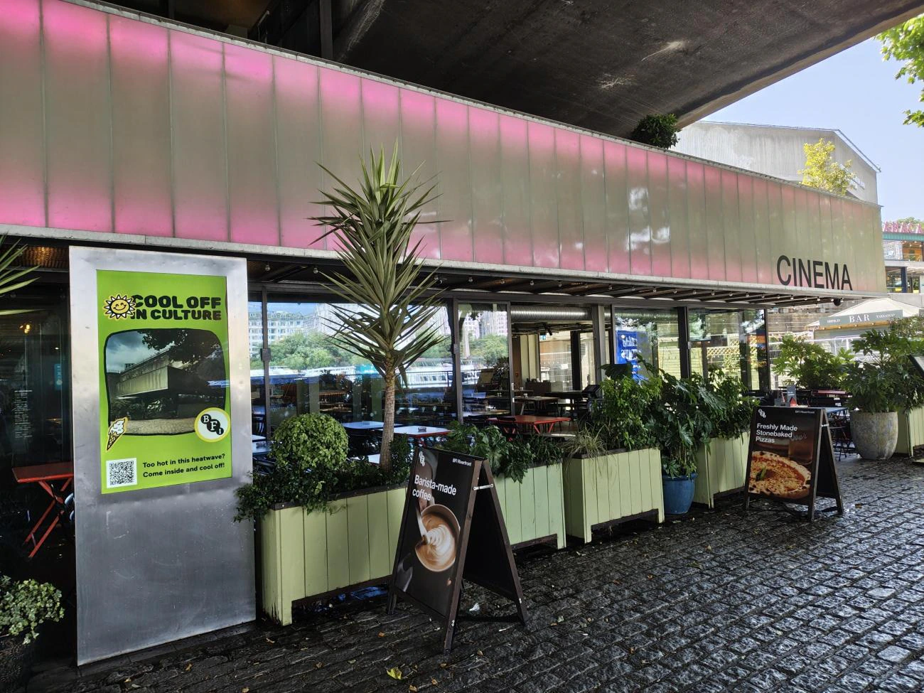 Outdoor view of a cinema with large glass windows, potted plants, and several sandwich boards promoting coffee and food specials. A “Cool Off in Culture” poster is displayed by the entrance.