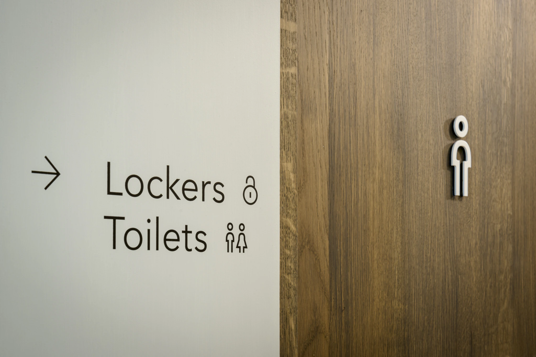 A sign on a wall points right to lockers and toilets, with icons for each. The adjacent wooden door displays a minimalist person icon, indicating a restroom.