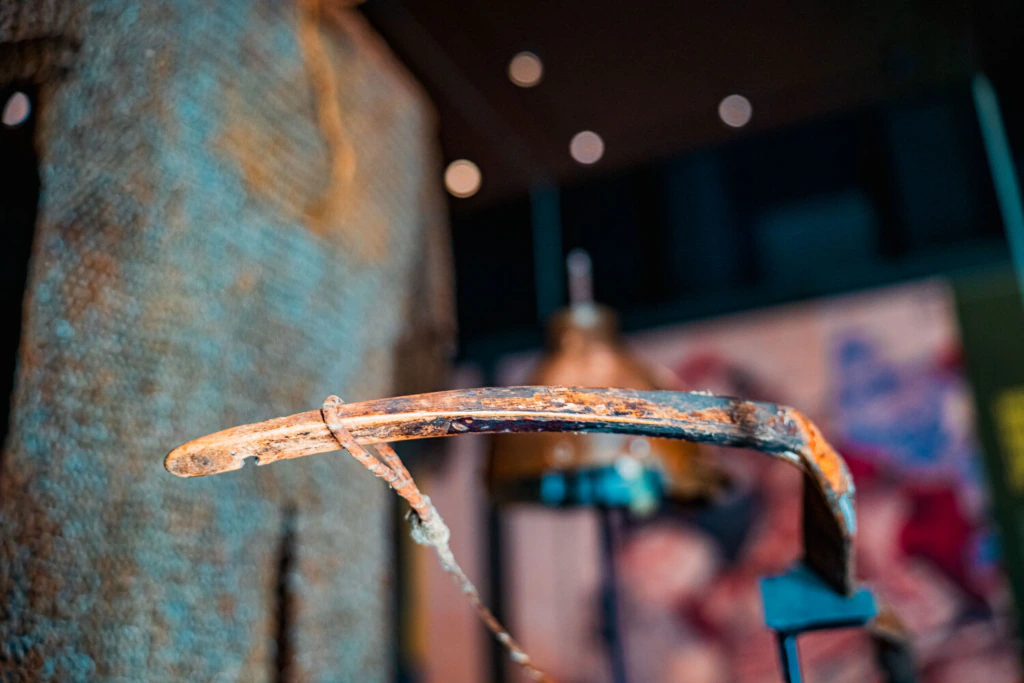 A close-up of an old, rusted farming tool or weapon displayed in a museum, with a blurred background of other artifacts and colorful displays.
