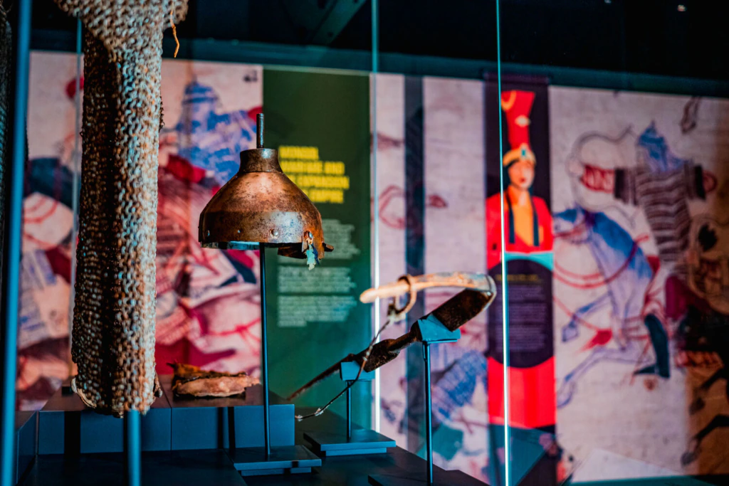 Ancient armor and helmet are displayed in a glass case at a museum, with blurred informational panels and illustrations of historical figures in the background.