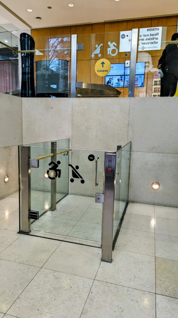 A small glass elevator platform for wheelchair access is built into the floor of a modern building, with accessibility symbols on the glass and a control panel on the side. Stairs and an entrance sign are visible above.