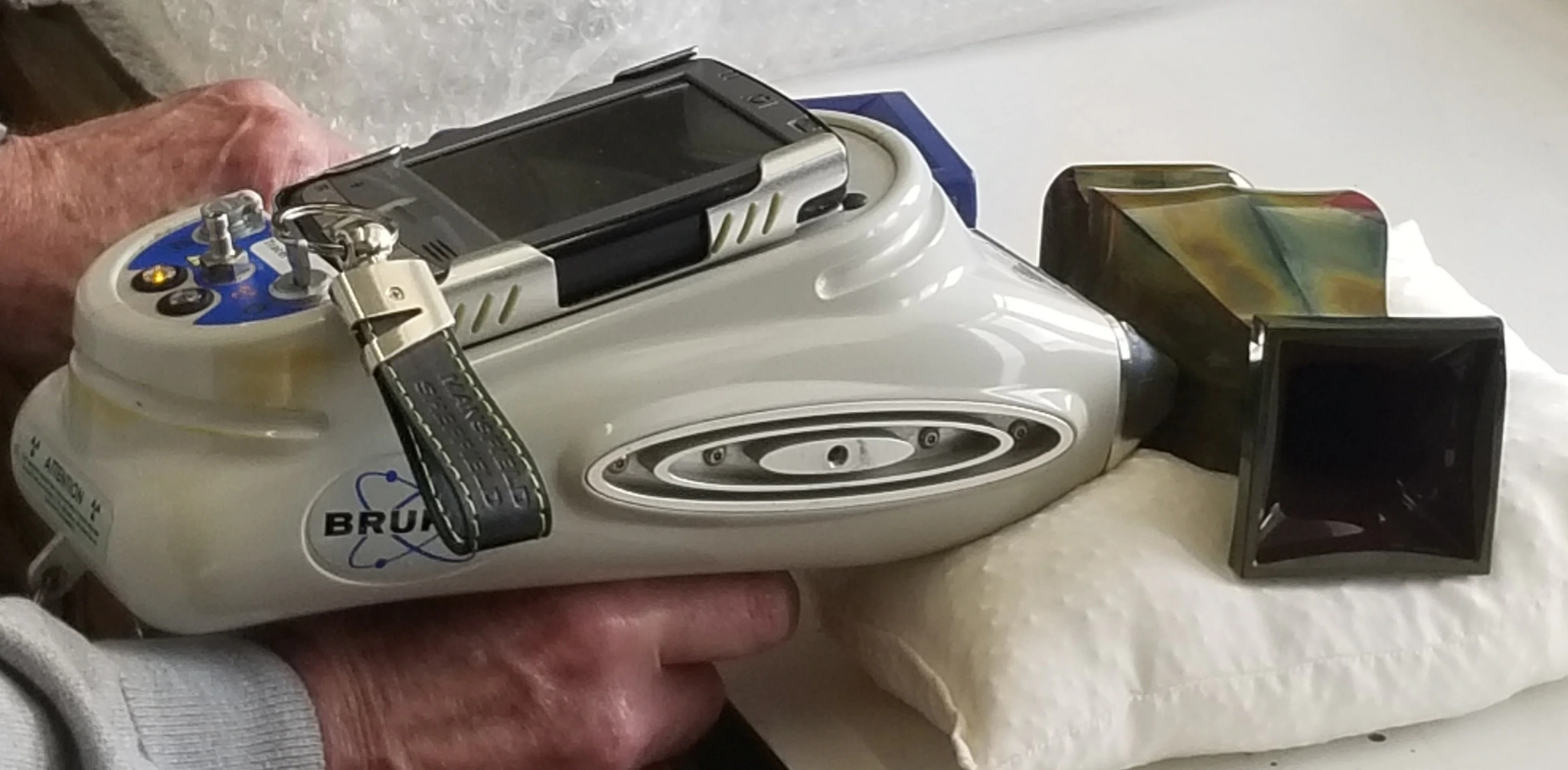 A person holds a handheld X-ray fluorescence analyzer, aimed at a metallic object placed on a cushion, likely for material or elemental analysis.