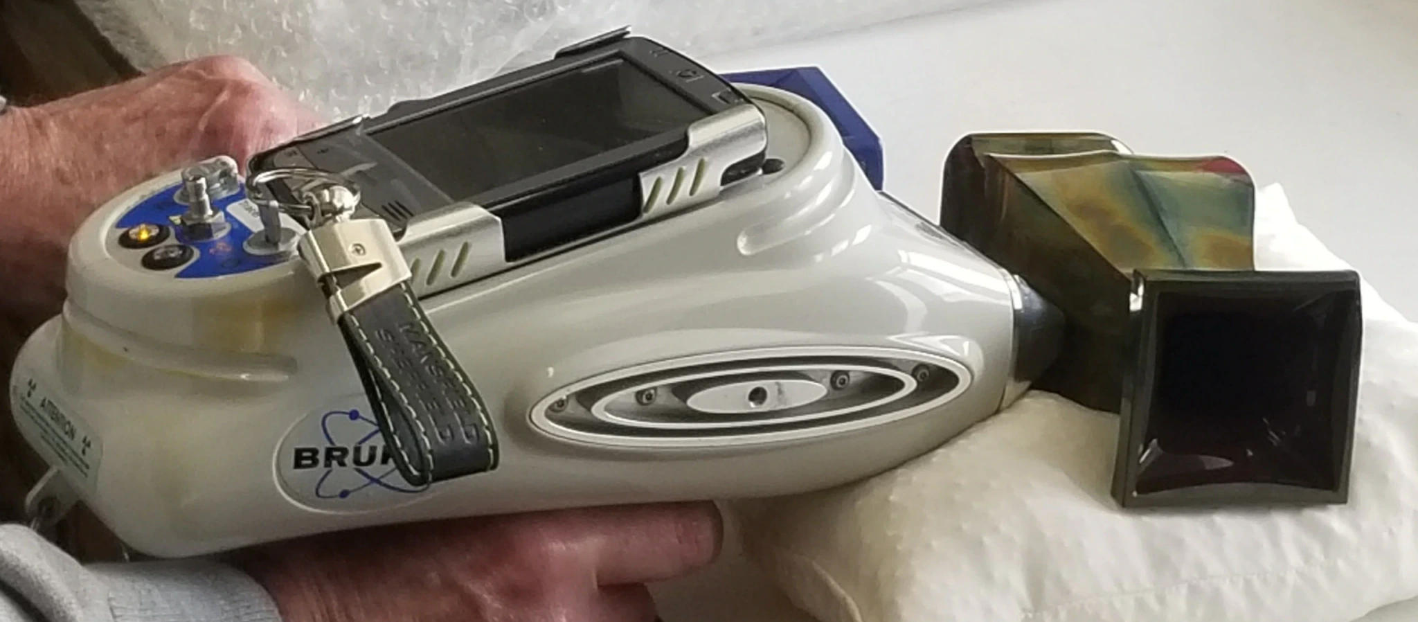 A person holds a handheld X-ray fluorescence analyzer, aimed at a metallic object placed on a cushion, likely for material or elemental analysis.