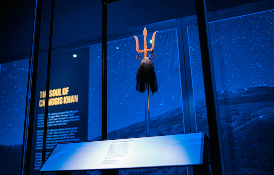 A ceremonial Mongolian black standard with a trident-shaped top and horsehair, displayed upright in a blue-lit museum exhibit titled The Soul of Chinggis Khan.