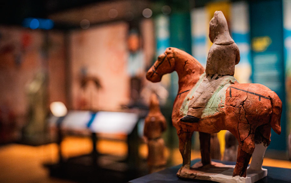 A cracked, ancient terracotta figurine of a rider on a horse is displayed in a museum, with soft, colorful lighting and blurred exhibits in the background.