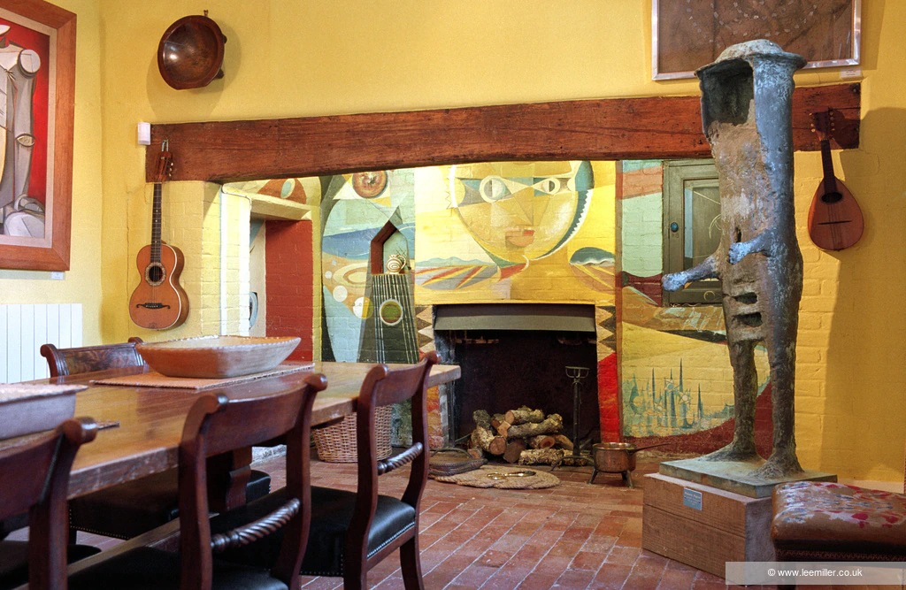 A colorful, eclectic room features a painted fireplace with abstract faces, wooden dining table and chairs, guitars hanging on yellow walls, a large modern sculpture, and various art pieces creating a vibrant, artistic atmosphere.