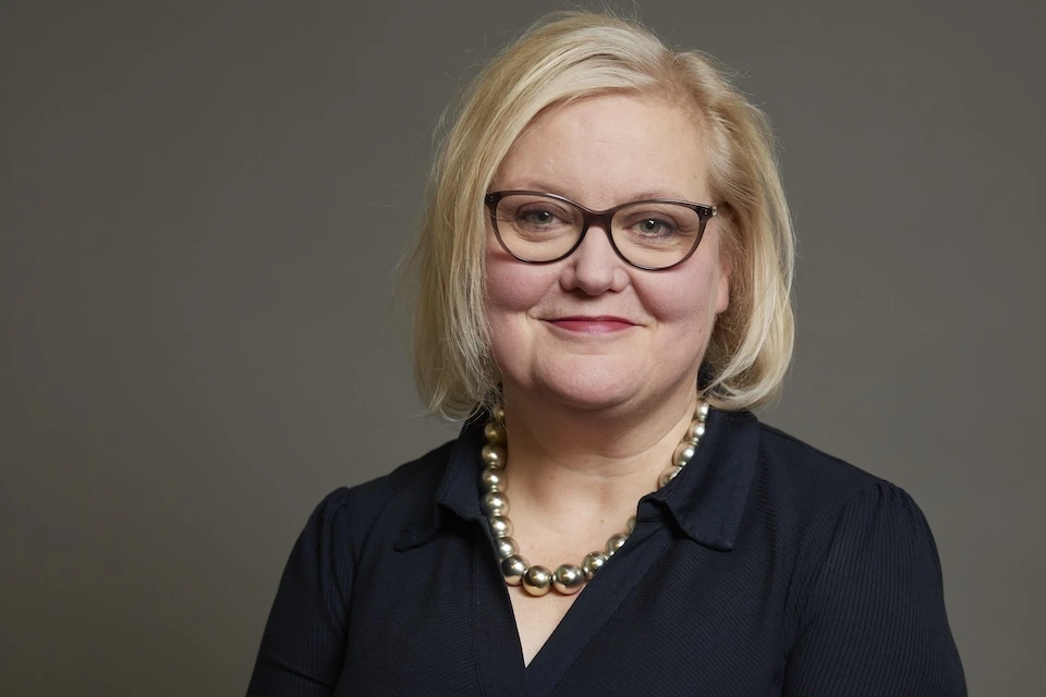 A woman with blonde hair, glasses, and a black top smiles at the camera. She is wearing a large pearl necklace and is posed in front of a plain gray background.