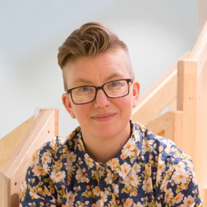 A person with short, styled hair and glasses smiles at the camera. They are wearing a blue floral shirt and sitting in front of a light-colored wooden staircase.