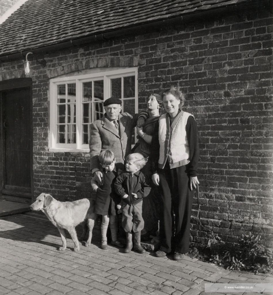 A black-and-white photo of a family standing outside a brick house. Two adults, three children, and a dog pose together. One adult holds a baby while the dog stands near a child in front. The group faces the camera, smiling.