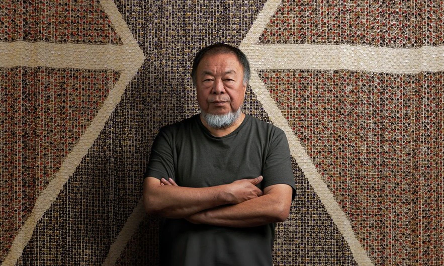 A man with a gray beard and mustache stands with arms crossed in front of a patterned tapestry with geometric designs. He wears a simple dark t-shirt and looks directly at the camera.