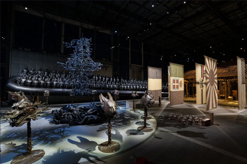 A dimly lit art exhibition features animal head sculptures on pedestals, a large black inflatable boat with human figures, hanging vertical banners, and other intricate installations under spotlights in a spacious hall.