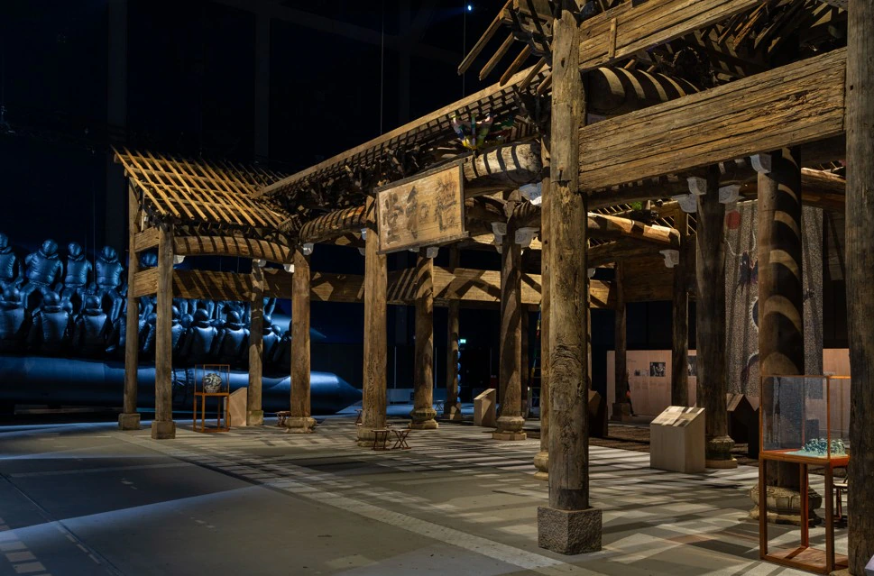A large wooden temple-like structure with exposed beams and columns is displayed in a dimly lit exhibition space, with glass cases and art objects nearby and a dark sculptural installation in the background.