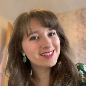 A woman with long brown hair and bangs smiles at the camera. She is wearing drop earrings and a green patterned top. The background is softly lit with neutral tones.