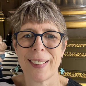 A smiling person with short gray hair, wearing round blue glasses and blue flower-shaped earrings, stands indoors in front of ornate gold and black decorations.