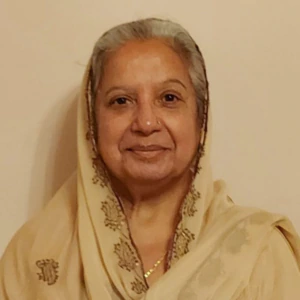 An older woman with gray hair smiles warmly. She is wearing a light beige traditional outfit with gold embroidery and a matching headscarf, standing against a plain cream-colored background.