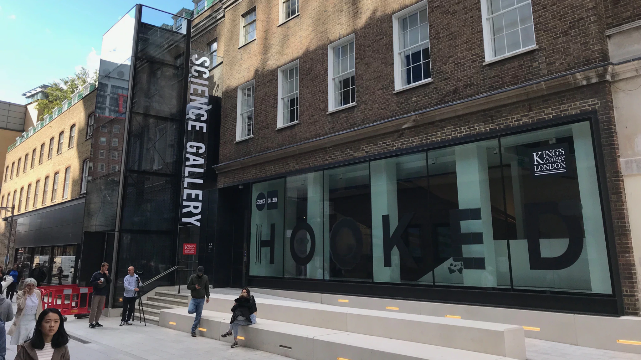 A brick building with large windows displays the words HOOKED and signs for Science Gallery and Kings College London. Several people walk or sit outside on a sunny day.