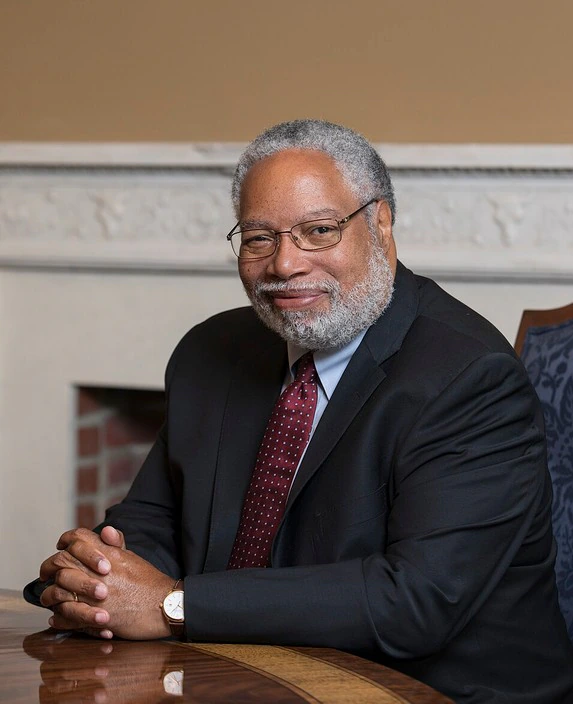 A distinguished older man with gray hair and a beard, wearing glasses, a dark suit, and a maroon tie, sits at a wooden table, smiling warmly with his hands clasped.