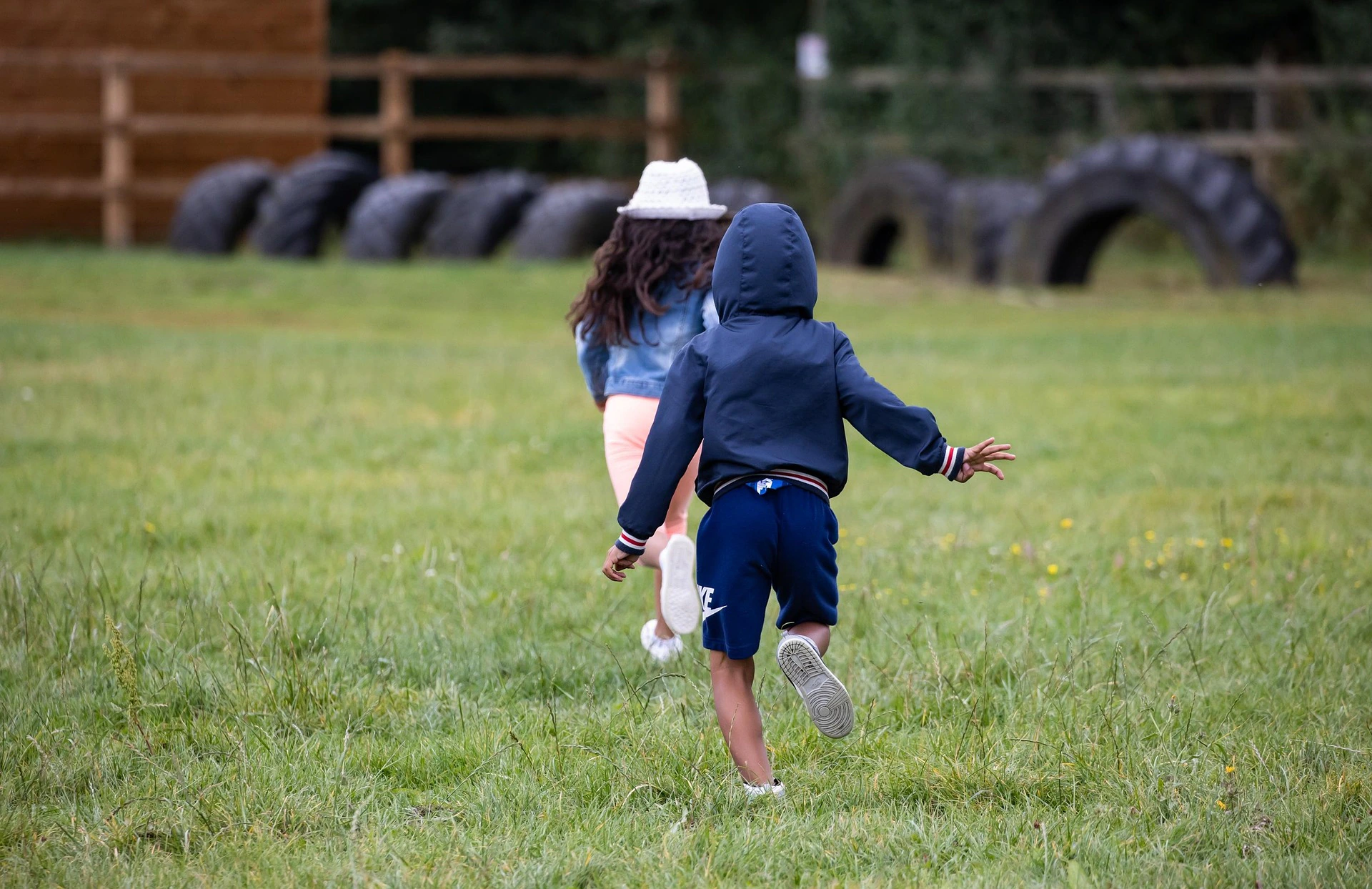 Two children, one wearing a white hat and the other a dark hoodie, run together across a grassy field with large tires and a wooden fence in the background. The children are seen from behind.