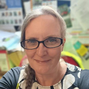 A woman with glasses and a braid smiles at the camera, wearing a patterned top. The background is brightly lit and cluttered with colorful papers and bulletin boards.