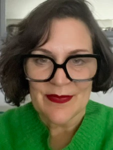 A woman with short dark hair, wearing oversized black glasses and a bright green sweater, smiles at the camera. She has bold red lipstick and a neutral background behind her.