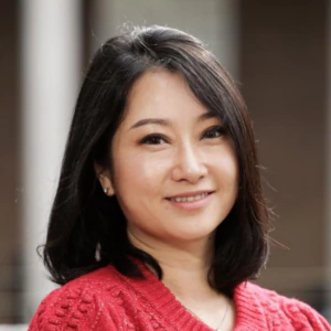 A woman with shoulder-length black hair wearing a textured red cardigan and black top smiles while standing outdoors in front of a building with white columns.