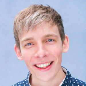 A young person with short, light brown hair and blue eyes smiles widely at the camera, wearing a navy blue shirt with white dots, against a light blue background.