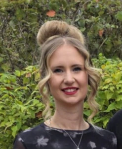 A woman with blonde hair styled in a high bun and loose curls stands outdoors in front of green foliage, wearing a black floral top and a silver necklace, smiling at the camera.