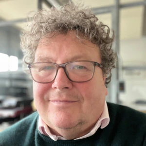 A man with curly hair, wearing glasses, a white collared shirt, and a dark green sweater, smiles slightly while sitting indoors in a bright room with large windows and industrial-style decor.