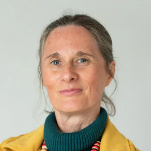 A woman with gray hair tied back, wearing a yellow jacket, teal turtleneck, and a chunky beaded necklace, stands with arms crossed against a plain light background.